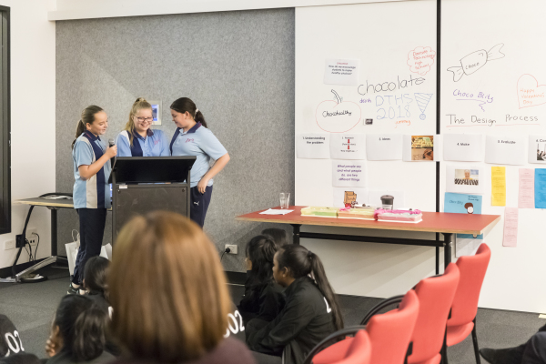 Three young people in school uniform presenting at a lectern, with a display in the background.